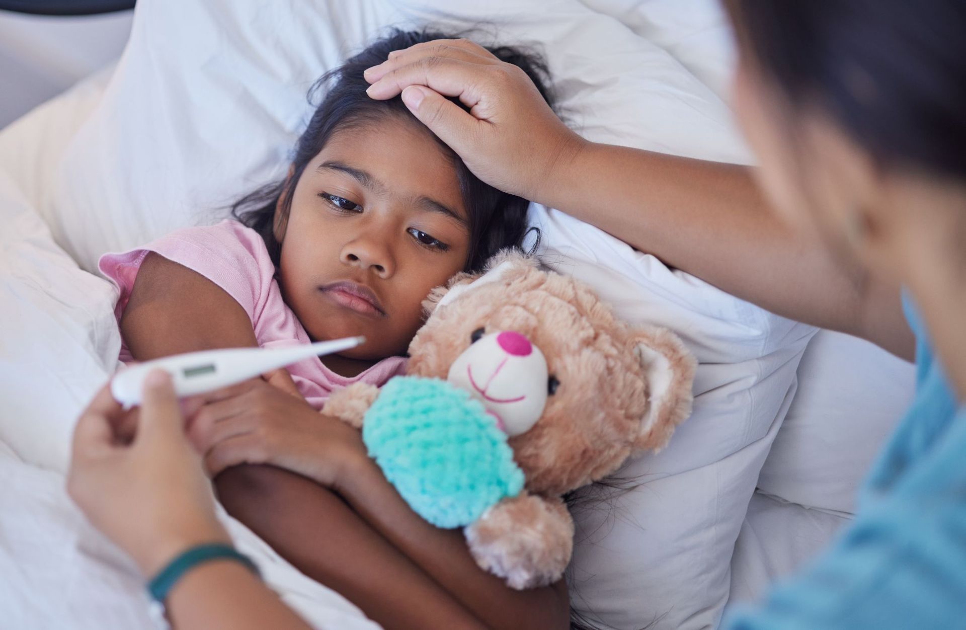 A little girl is laying in bed with a teddy bear and a thermometer in her mouth.