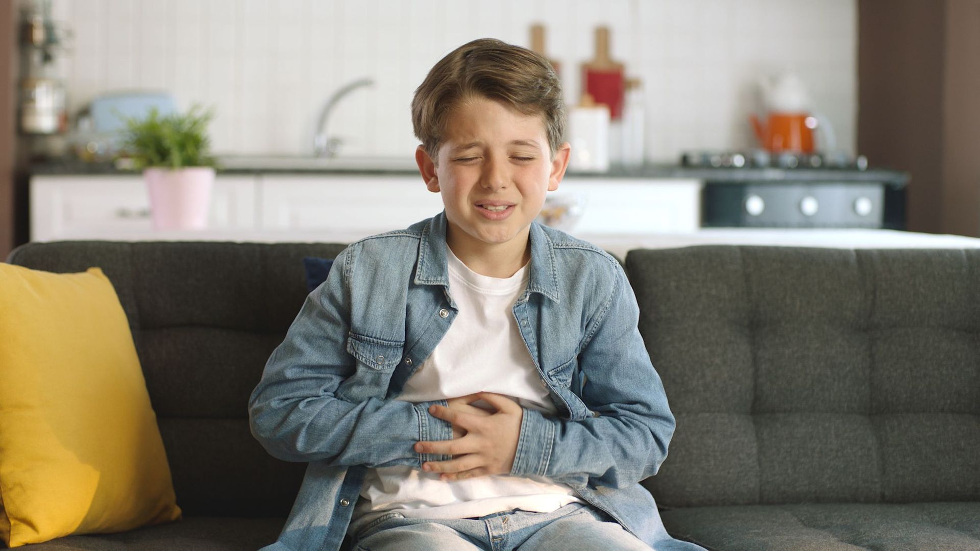 A young boy is sitting on a couch holding his stomach.