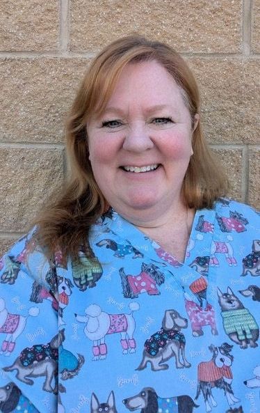 A woman wearing a blue scrub top with dogs on it is smiling in front of a brick wall.